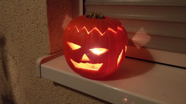 Geschnitzte Kürbislaterne mit gezacktem Deckel und grinsendem Gesicht, innen leuchtet eine Kerze, auf einem Fensterbrett