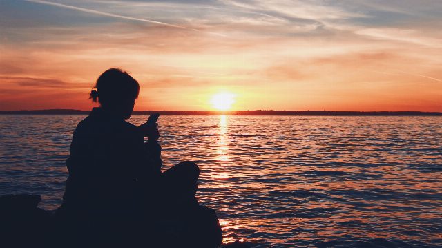 Person sitzt bei Sonnenuntergang am Wasser und schaut auf ihr Smartphone, nur als Silhouette zu sehen