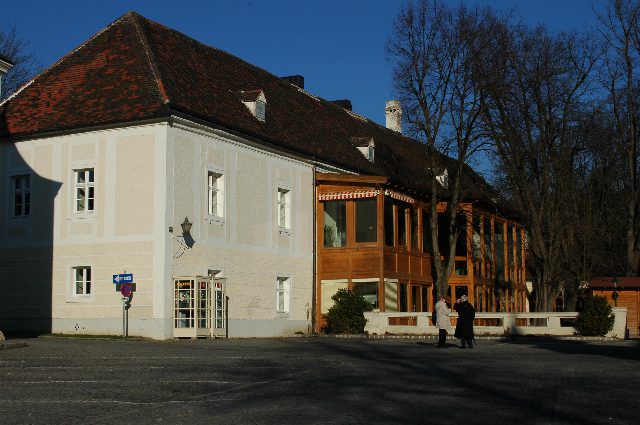 Der Klostergasthof Heiligenkreuz: ein langgestrecktes weißes Haus mit Ziegeldach und verglastem Holzvorbau, davor ein gepflasterter Platz mit zwei Spaziergängern