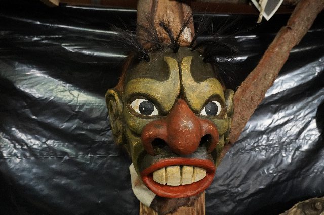 Geschnitzte Holzmaske mit grünlichem Gesicht, roter Nase, roten Lippen, großen Zähnen und schwarzen Haarbüscheln, vor dunkler Folie