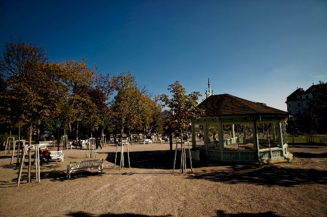Herbstlich gefärbte Bäume und ein achteckiger Pavillon mit Holzdach auf einem sonnigen Kiesplatz im Kurpark Baden, daneben Parkbänke
