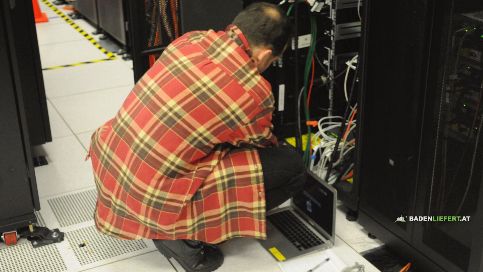 Ein Techniker im karierten Hemd hockt mit einem Laptop vor einem geöffneten Serverschrank in einem Rechenzentrum und arbeitet an der Verkabelung