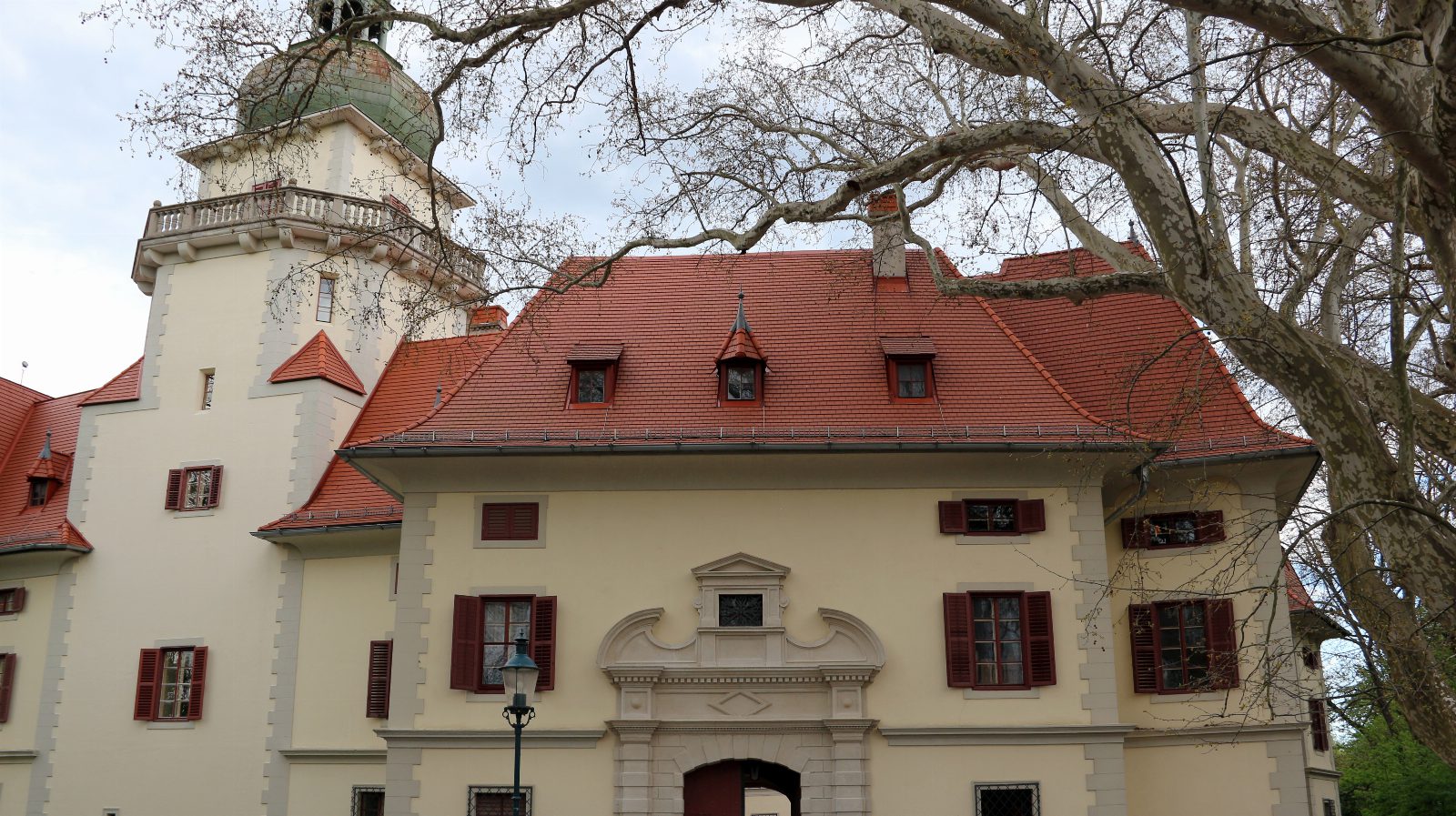 Hellgelbes Schloss mit rotem Ziegeldach, Torbogen und Turm mit grüner Kuppel, davor die Äste einer großen kahlen Platane