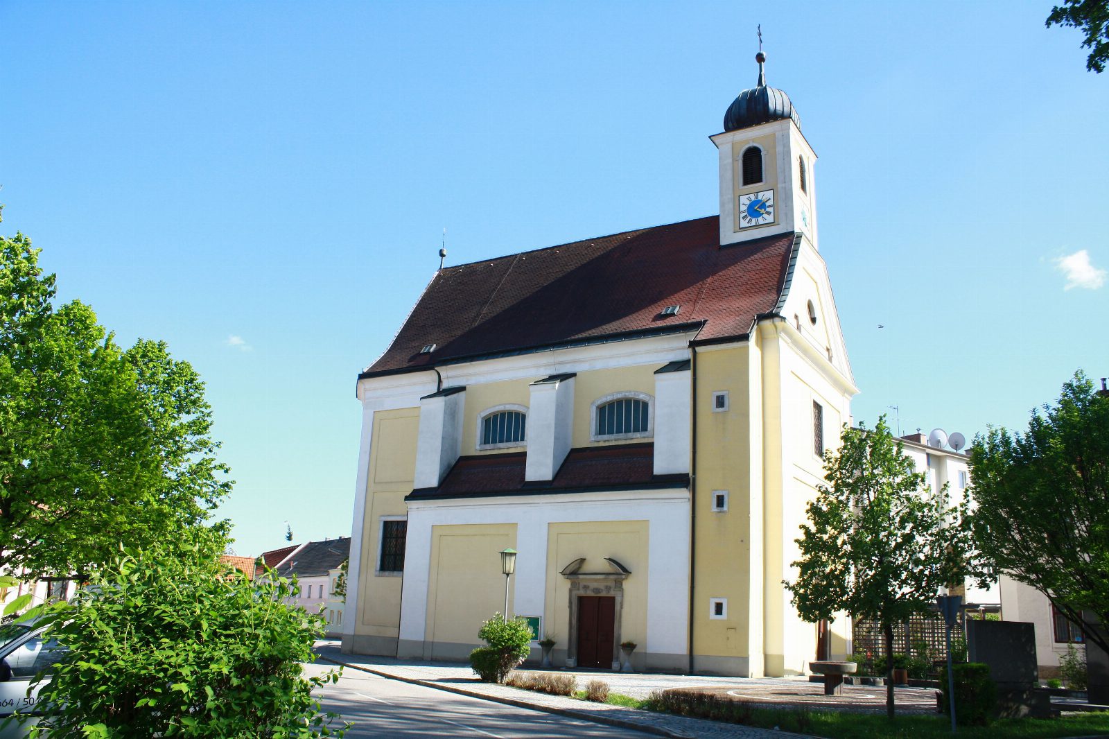 Gelb-weiße Barockkirche mit Zwiebelturm und Uhr unter blauem Himmel, davor eine Straße und grüne Bäume