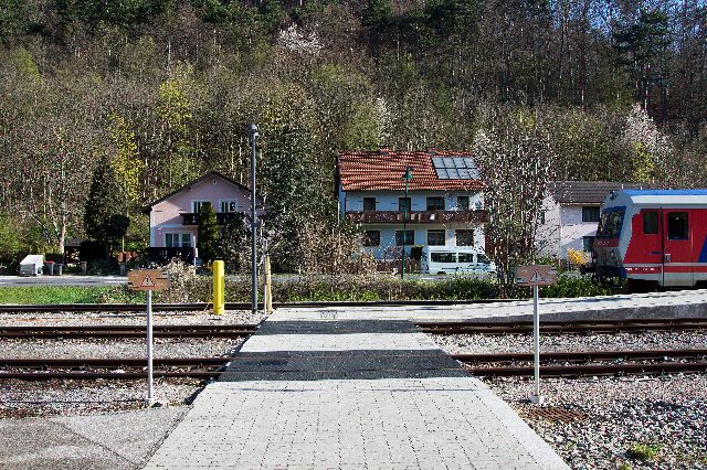Bahnübergang am Bahnhof Weissenbach-Neuhaus, rechts ein rot-weißer Triebwagen der ÖBB, dahinter Wohnhäuser und bewaldeter Hang